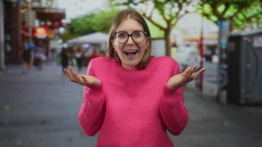 Geniş bir gülümseme ve kalabalık bir şehir caddesinde açık palmiyeler gösteren genç sarışın kadın ağaçlar ve kafelerle dolu; neşe.