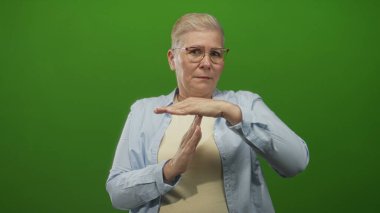 Yeşil bir stüdyo ortamında, gözlüklü ve sıradan gömlekli, kendine güveni tam bir jest yapan elleri olan bir kadın..