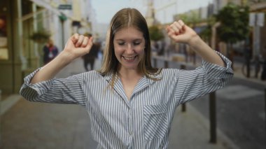 Elleri sımsıkı bağlanmış, sokakta gülümseyen, mağazaların önünde çizgili tişört giyen ve şehir binalarının altında sokak lambaları taşıyan bir kadın..