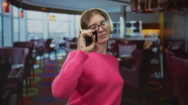 Kulaklık takan genç sarışın kadın akıllı telefonu dudaklarına tutarak canlı bir restoranda şarkı söylüyor..
