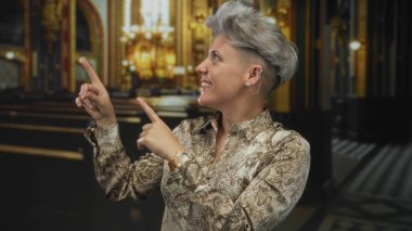 Kısa saçlı, gülümseyen ve parmakları loş kilisedeki ahşap sıraların üzerinde sunağı gösteren bir kadın..