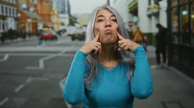 Yoğun bir şehir sokağında, arka planda arabaları ve yayaları olan sokak hayatını gözler önüne seren, neşeli bir ifade sergileyen gri saçlı son sınıf kadını..