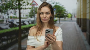 Genç beyaz kadın işlek şehir caddesinde iki eli ve parşömen ekranı olan akıllı telefonu tutuyor; dijital bağlantı.