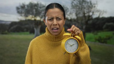 Yeşil bir parkta elinde sarı alarmlı saat tutan bir kadın bulutlu bir günde arka planda yemyeşil ağaçlarla hayal kırıklığını ifade ediyor..