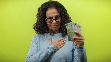 Arjantinli banknotları elinde tutan orta yaşlı bir kadın. Bir eli göğsünde, izole edilmiş sarı arka planda mali sorumluluk ve gururu yansıtan bir kadın..