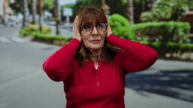 Kırmızı kazaklı yaşlı İspanyol kadın yeşil ve güneş ışığıyla çevrili sessiz şehir caddesinde kulaklarını kapatıyor. Stres ve gürültü hassasiyetini ifade ediyor..