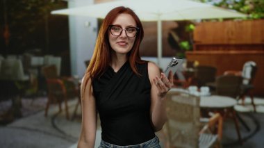 Terasta gülümseyen bir kadın dışarıdaki kafede akıllı telefon tutuyor. Etrafı sandalyeler ve masalarla çevrili ve sokak manzarasını güzelleştiriyor..