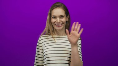 Genç beyaz kadın mor stüdyo ortamında el sallıyor parlak ışıklandırma ve sıradan çizgili gömlek görünür; mutluluk sıcaklığını karşıla.