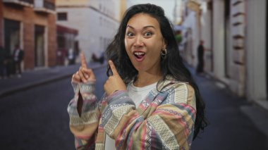 Bir kadın açık ağızlı, açık parmaklı, desenli ceket giyen, jest yapan ve gülümseyen birini işaret ediyor..