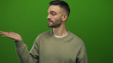 Latin kökenli sakallı genç adam elini uzatıyor ve bej bir süveter giymiş, koyu yeşil bir arka plana karşı derin düşüncelere dalmış bir ifade sergiliyor..