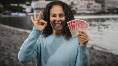 İsrail şekellerini tutarken gülümseyen bir kadın deniz kenarında arka planda bulanık su olan bir plajda iyi el sallıyor..