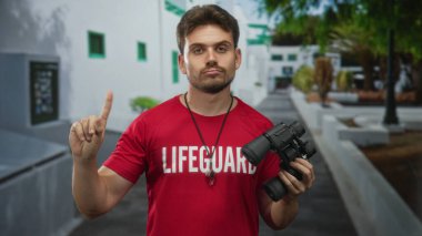 Can kurtaran cankurtaranı, can kurtaran baskısı ve ıslık kolyeli kırmızı t-shirt giyer. Binada dürbün tutarken parmağı gösterir..