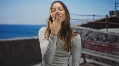 Kadın, deniz kenarındaki bir bankta el pençe dudağa bir öpücük savuruyor, beyaz bluz giyiyor ve gülümsüyor; neşeli bir sevgi..