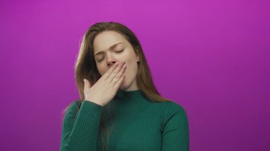 Yeşil kazaklı genç kadın başını eğiyor ve elleri mor stüdyo ortamında ağzını kapatıyor..