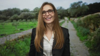 Genç bir kadın, yapraklı bir park patikasında dururken siyah ceket ve beyaz gömlek içinde aşağılanmış gözler ve sarkık dudaklarla kaşlarını çatar;.