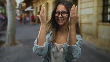 Kadın, taş binaların yakınındaki bulanık bir şehir caddesinde dururken sarı çivi ve yüzükleri gösteren ellerle yüzünü kapatıyor..