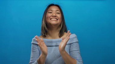 İzole edilmiş mavi bir arka plan duvarına karşı coşkuyla alkışlayan bir kadın, coşkulu bir gülümsemeyle neşe ve pozitiflik sergiliyor, mutluluk ve başarıyı ifade etmek için mükemmel..