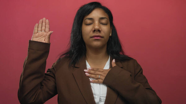 African american woman raises hand and touches chest with eyes closed in pink studio; pledge sincerity.