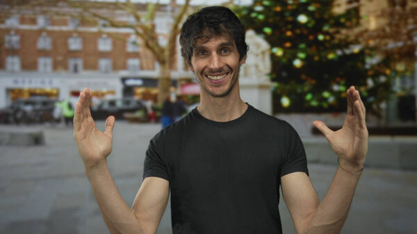 Man spreads bare forearms and open palms toward camera with smiling face on street lined with blurred lights overhead; joy.