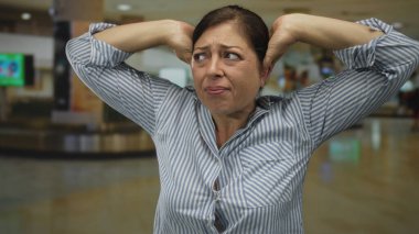 Orta yaşlı mavi çizgili gömlekli kadın havaalanı terminalinde kulaklarını gergin bir ifadeyle kapatıyor..