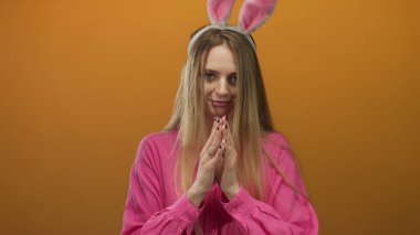 Tavşan kulaklı kadın turuncu bir arka plana gülümsüyor pembe bir gömlek giyiyor, neşeli bir paskalya temasını canlı bir şekilde yakalıyor..