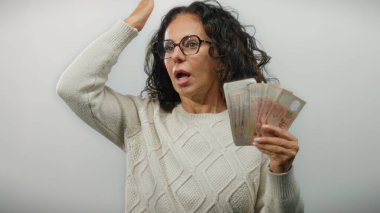 Orta yaşlı bir kadın, beyaz bir duvarın karşısında para sergiliyor. Şaşırdığını ve finansal açıdan başarılı olduğunu söylüyor..