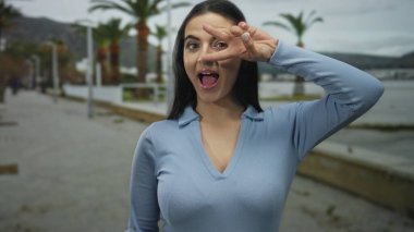 Genç İspanyol kadın deniz kenarında bulutlu gökyüzünün altında palmiye ağaçlarıyla zafer jesti yapıyor. Mavi kıyafetiyle neşe ve pozitifliğini ifade ediyor..