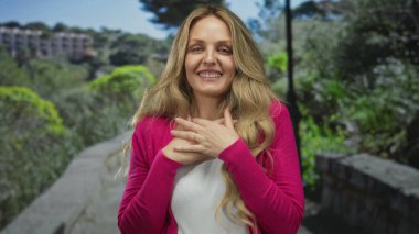 Parkta mutlu bir şekilde gülümseyen bir kadın güneşli bir günde pembe bir kazak giyiyor. Etrafında canlı yeşillikler ve ağaçlar var..