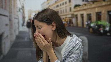 Çizgili tişört giyen genç İspanyol kadın işlek bir şehir caddesinde iki eliyle ağzını kapatıyor..