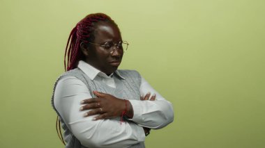 Saçları örgülü, gözlüklü ve gri yelekli Afro-Amerikalı kadın kollarını izole edilmiş sarı arka planda birleştiriyor, özgüven ve tefekkür iletiyor..