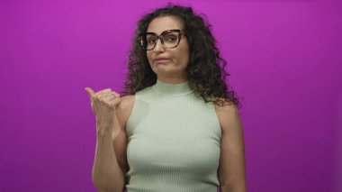 Stüdyoda yeşil balıkçı yaka giyen, mor arka planda başparmağı yana gösteren bir kadın..