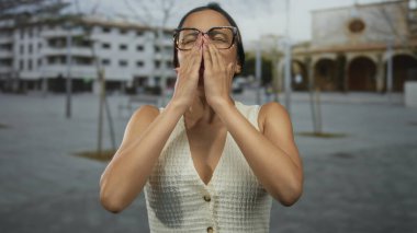 İspanyol kadın gözlüklü ve beyaz bluzlu bir kadın şehir sokağında duruyor, yüzünü ellerle örtüyor, duygularını modern bir açık hava ortamında aktarıyor..