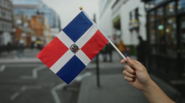 El ele tutuşan Dominik Cumhuriyeti bayrağı, şehir arka planına karşı canlı renkler sergiliyor, ulusal gurur ve kültürel kimliği sembolize ediyor..