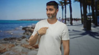 Erkek, beyaz t-shirt ve bilezik giyen, palmiye ağaçları, kayalık kıyılar ve gezinti güvertesi ile kaplı bir sahil caddesinde parmak göğse işaret eder..