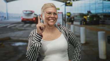 Kadın, havaalanı terminalinde geniş bir gülümseme içinde, çizgili gömlek ve gözlük takarken yukarı doğru işaret ediyor; güven yolculuğu ipuçları.