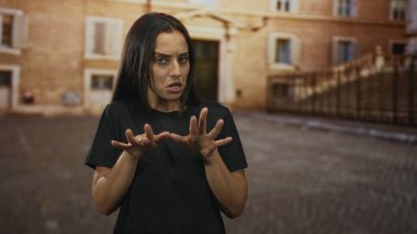 Elleri önde uzanan ve parmakları taştan bir bina tarafından eski bir kasaba caddesinde yayılmış bir kadın, gergin jestler yapıyor; sıkıntı.