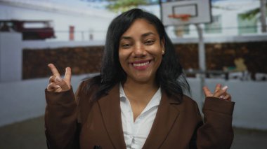 Kadın açık hava basketbol sahasında beyaz gömlek giyerek zafer işareti gösterirken geniş bir gülümseme sergiliyor; barış umudu birliği.