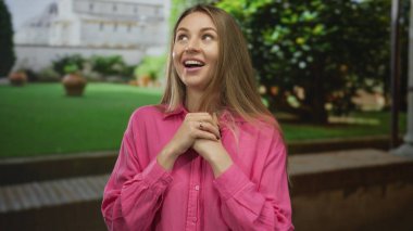 Dışarıda pembe tişörtlü bir kadın, arka planda yeşil bahçeli ve tarihi binalı bir sevinç ifadesi sergiliyor, mutluluğu, gençliği ve doğanın güzelliğini vurguluyor..