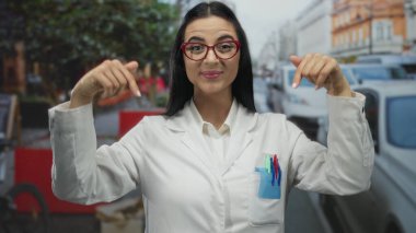 İşlek bir caddeyi işaret eden gözlüklü İspanyol kadın bilim adamı beyaz laboratuvar önlüğü giyiyor açık havada canlı bir şehir ortamında kalemler sergiliyor.