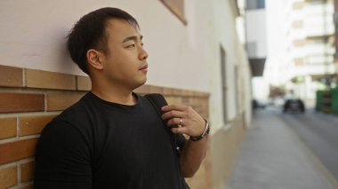 Şehirdeki bir duvarın önünde duran genç adam, şehirde düşünceli bir yüz ifadesi sergiliyor..