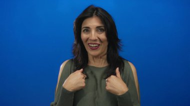 Orta yaşlı İspanyol bir kadın, parlak bir gülümseme ile mavi bir stüdyo duvarına karşı kendini işaret ediyor..