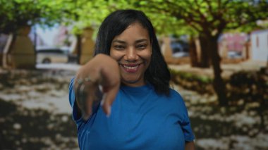 Mavi gömlekli kadın geniş bir gülümseme ve parktaki yeşil ağaçlar arasında parmak işareti; dostça bir davet.