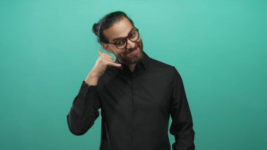 Gözlüklü ve sakallı genç adam turkuaz stüdyoda jest yapıyor. Siyah gömlek giyiyor ve gülümsüyor..