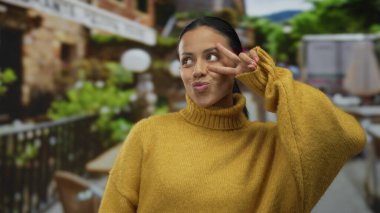 Terasta hardal süveter giymiş bir kadın arka planda bulanık bir açık hava restoranıyla barış işareti sergiliyor. Büyüleyici bir İspanyol atmosferi olduğunu gösteriyor..
