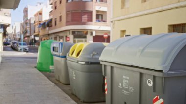 Gri ve yeşil geri dönüşüm kutuları ve güneşli bir şehir caddesinde sığ bir bokeh 'te yumuşak odaklı binalar ve kaldırımlarla yakalanan park edilmiş arabalar; arkaplan telif alanı.