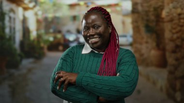 Çapraz kollu Afro-Amerikalı bir kadın açık hava sokak sahnesinde güvenle gülümsüyor yeşil bir kazak giyiyor ve canlı kızıl saçlar sergiliyor..