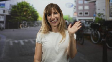 Kadın beyaz tişörtle gülümserken el sallıyor, park halindeki bisikletlerin yanında duruyor ağaç ve bina cephesi; neşeli bir selamlama.