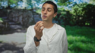 Işıl ışıl, yemyeşil bir parkta jest yapan genç bir İspanyol adam, günlük beyaz gömlek ve saat giyiyor, canlı doğayla çevrili ve gevşemiş rahatlama tavrını canlandırıyor..