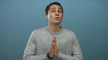 Genç İspanyol bir adam mavi arka plan için gözleri kapalı dua ediyor, umut ve maneviyatını ifade ediyor..