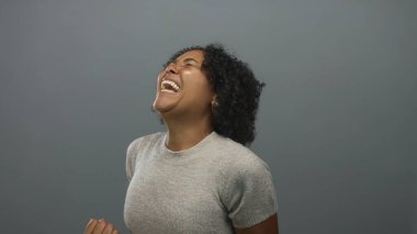 Genç Afrikalı Amerikalı kadın gri bir stüdyoda yumruklarını sıkar ve başını geriye atar; heyecan.